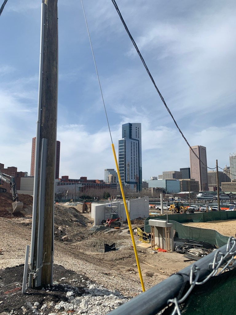 A construction site with lots of gravel, dirt, holes, and diggers. Tall buildings are in the background.
