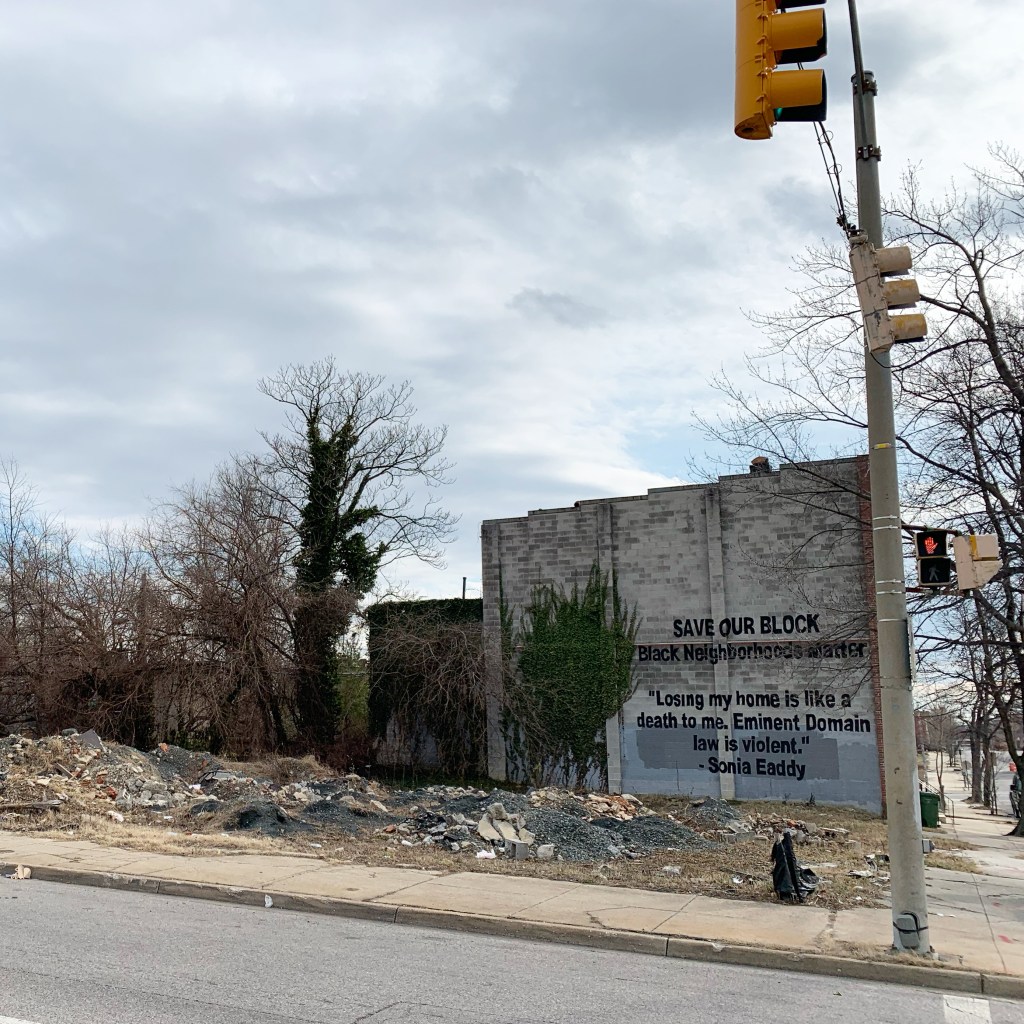 A picture of the side of a house with a mural that reads: SAVE OUR BLOCK. Black Neighborhoods Matter. "Losing my home is like a death to me. Eminent Domain law is violent." --Sonia Eaddy

There is rubble in the foreground from a house that has been torn down next to it.
