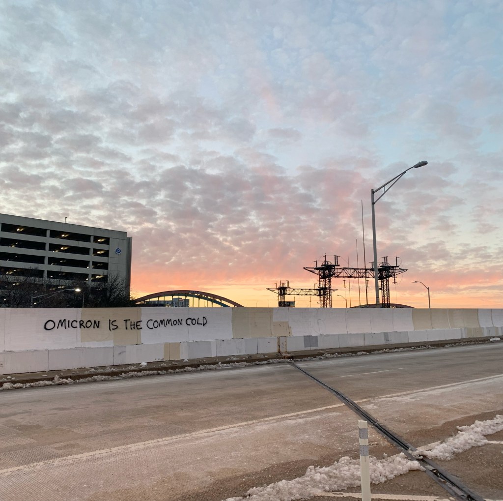 Picture of a cloudy sky at sunset, light orange and pink at the bottom and stretching up to clouds and light blue sky. There is graffiti on the bridge in the foreground that reads, "OMICRON IS THE COMMON COLD."