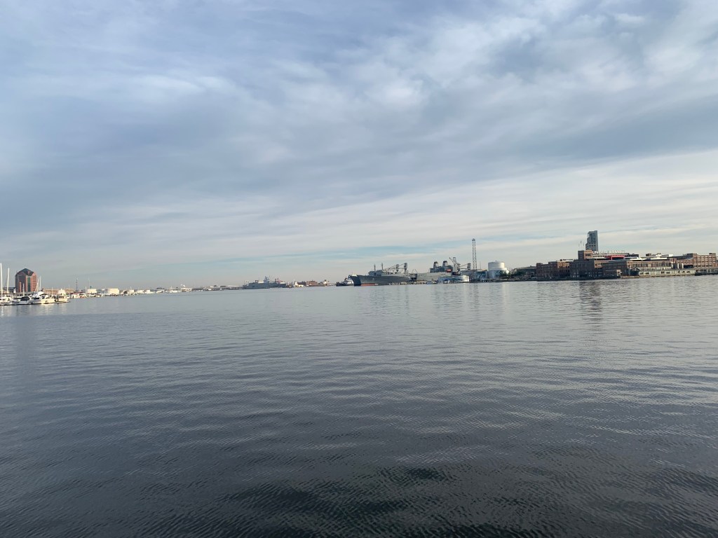 The bottom half of the picture is blue-grey water and the top half is gray clouds with just a sliver of blue in the foreground. The picture is divided by a line of buildings across Baltimore's harbor.
