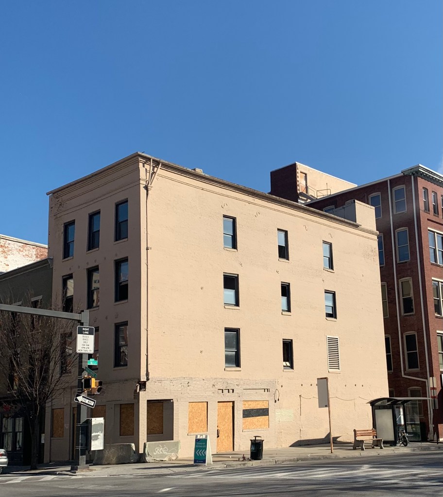 Picture of a four story beige brick building with boarded-up windows on the first floor against a bright blue sky.