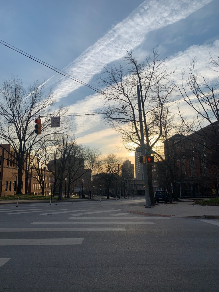Picture looking down the street at an intersection. The sun is coming up about midway from the horizon. The sky is streaked with clouds and new light, and there are leafless trees against it.