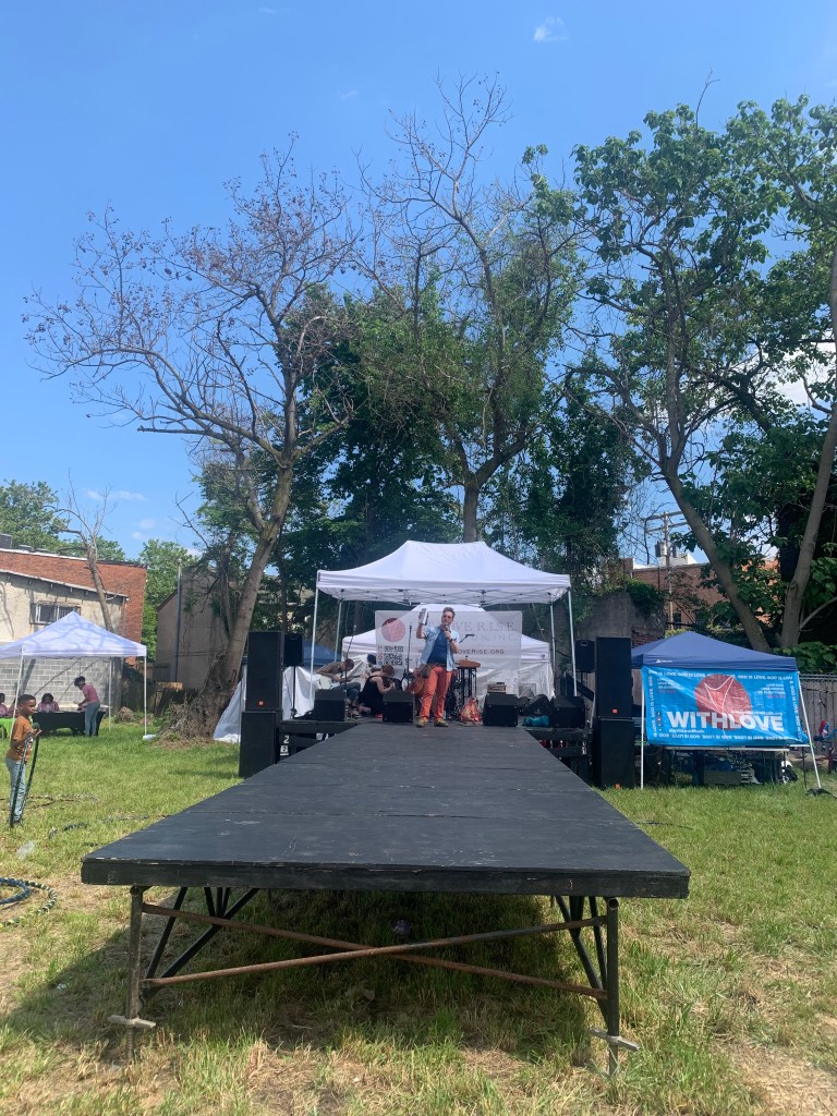Picture of a white man in salmon pink pants, a blue t-shirt, and a blue button up over the top of it. He's standing at the end of a long platform set up on grass that leads to a stage where string musicians are setting up. There are tents on either side, and a kid standing with a hula hoop. The sky is bright blue with just a few puffy white clouds.