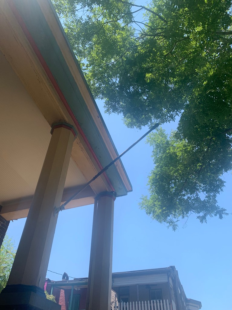 Looking up at a blue sky and porch roof. There is an empty flag pole on the house. There is a green tree at the right of the picture.