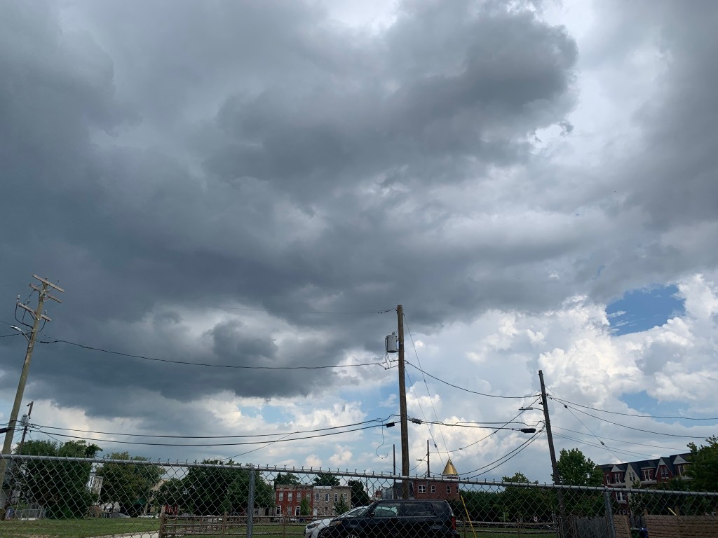 A chain link fence is in the foreground, and the bulk of the picture is of the sky. It is filled with gray clouds, with some blue sky peeking out.